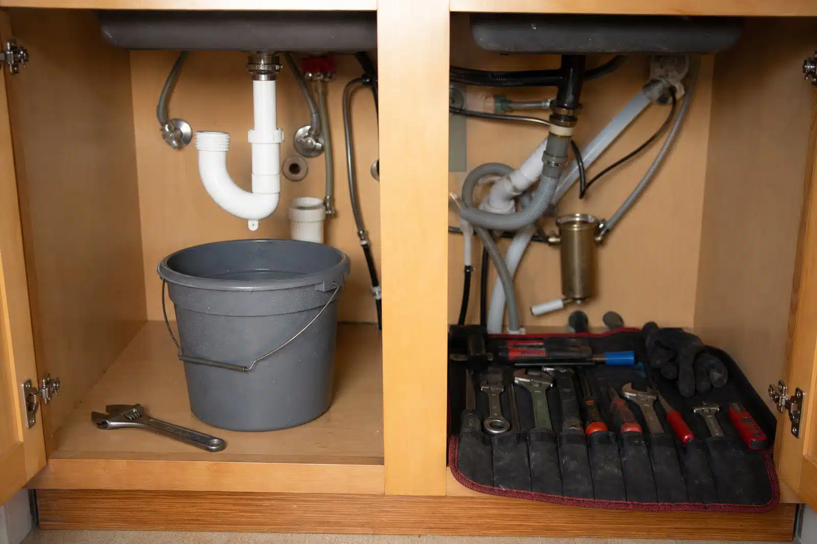 An open kitchen sink cabinet with a homeowner's single wrench and a bucket under a half-disassembled P-trap on one side and a plumber's organised tool roll on the other.