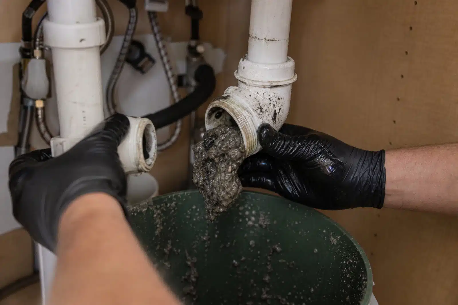 Gloved hands opening a P-trap under a kitchen sink over a bucket, thick grey grease sludge visible blocking the trap.